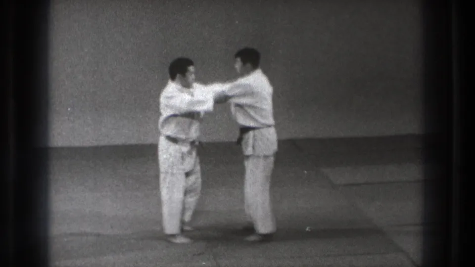 In 1971 Tokyo footage, two karate students in white gi stand facing each other during partner practice on a matted floor.