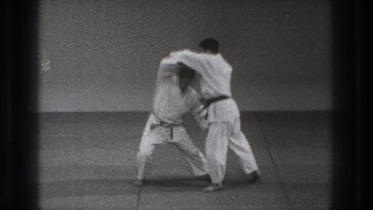 Two karate students continue the technique, one crouching low while the other maintains a close upper-body hold in the dojo-like room.