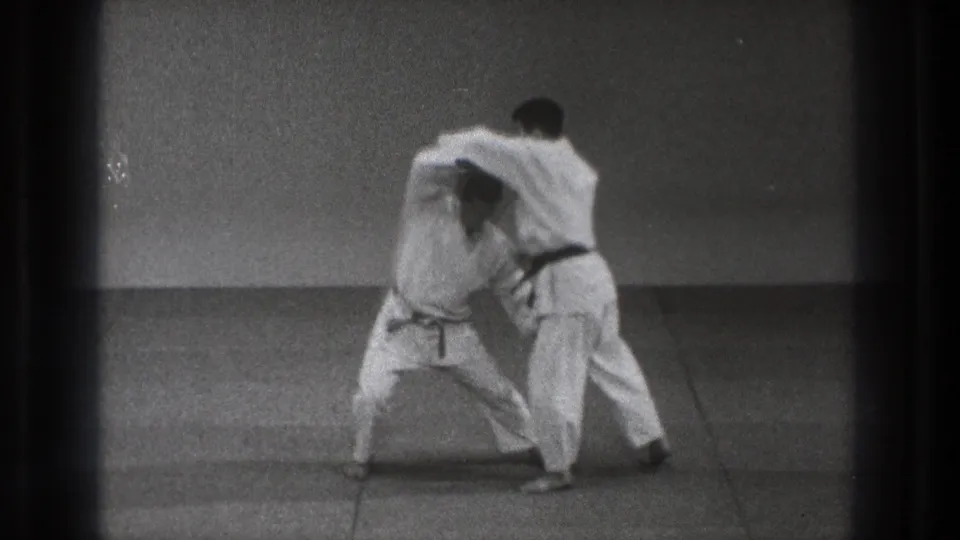 Two karate students continue the technique, one crouching low while the other maintains a close upper-body hold in the dojo-like room.