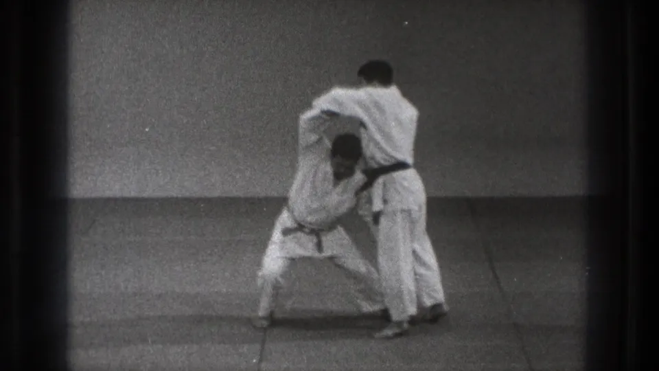 Two martial artists in white gi and black belts practice a close hold on a simple mat in Tokyo, Japan, 1971 footage.