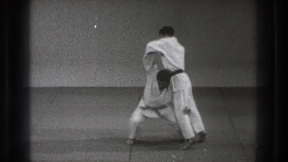 In 1971 Tokyo karate footage, one black-belt practitioner braces while controlling his partner during a throw setup.