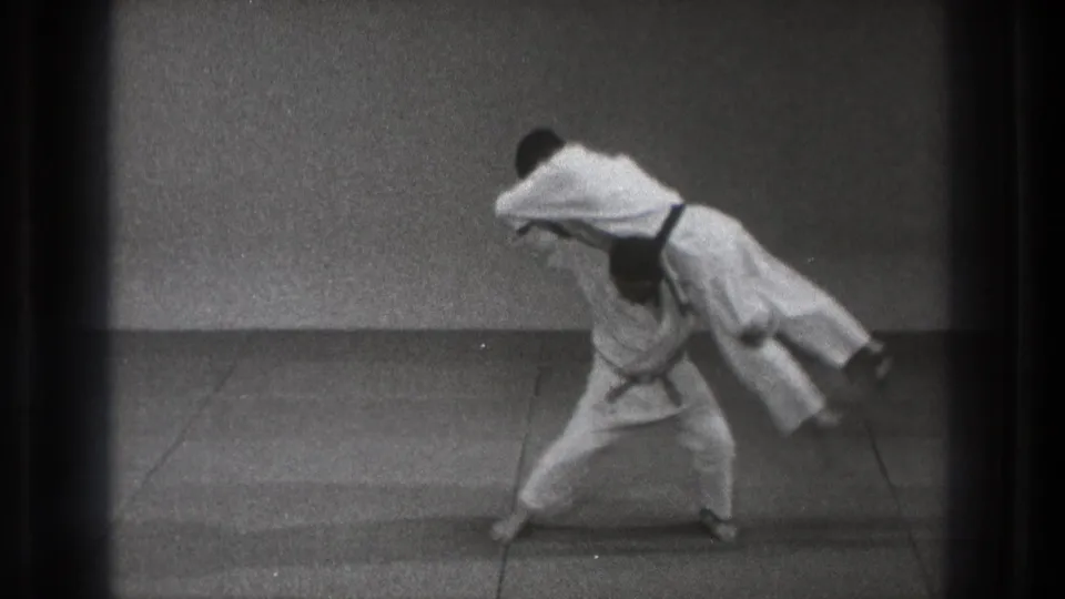 The throw reaches its peak as one martial artist lifts his partner nearly horizontal on a plain mat in 1971 Tokyo footage.