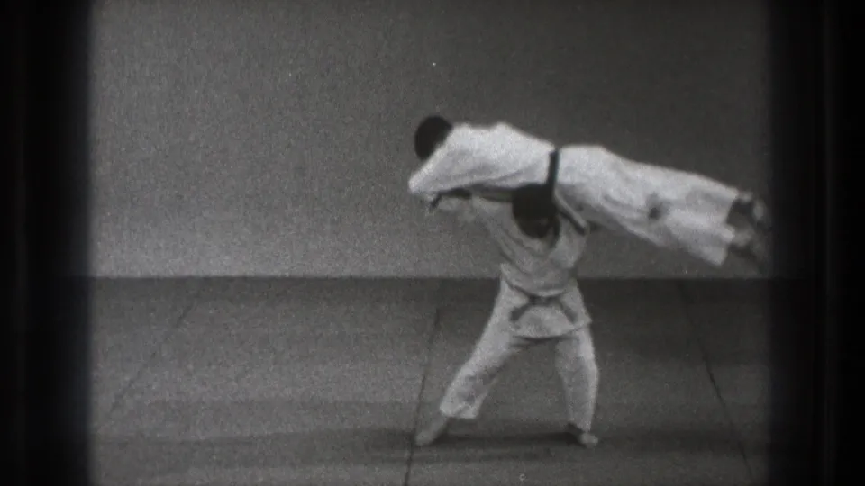In 1971 Tokyo footage, one martial artist in a white gi lifts another across his shoulders during a demonstration.