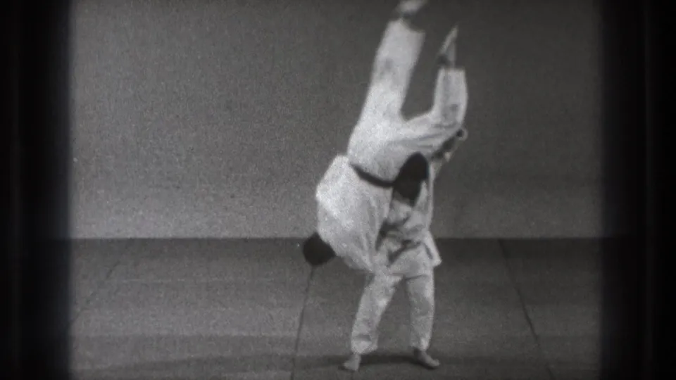 In supplied 1971 Tokyo footage, one man in a white judo uniform lifts another high during a throw on a plain indoor mat.