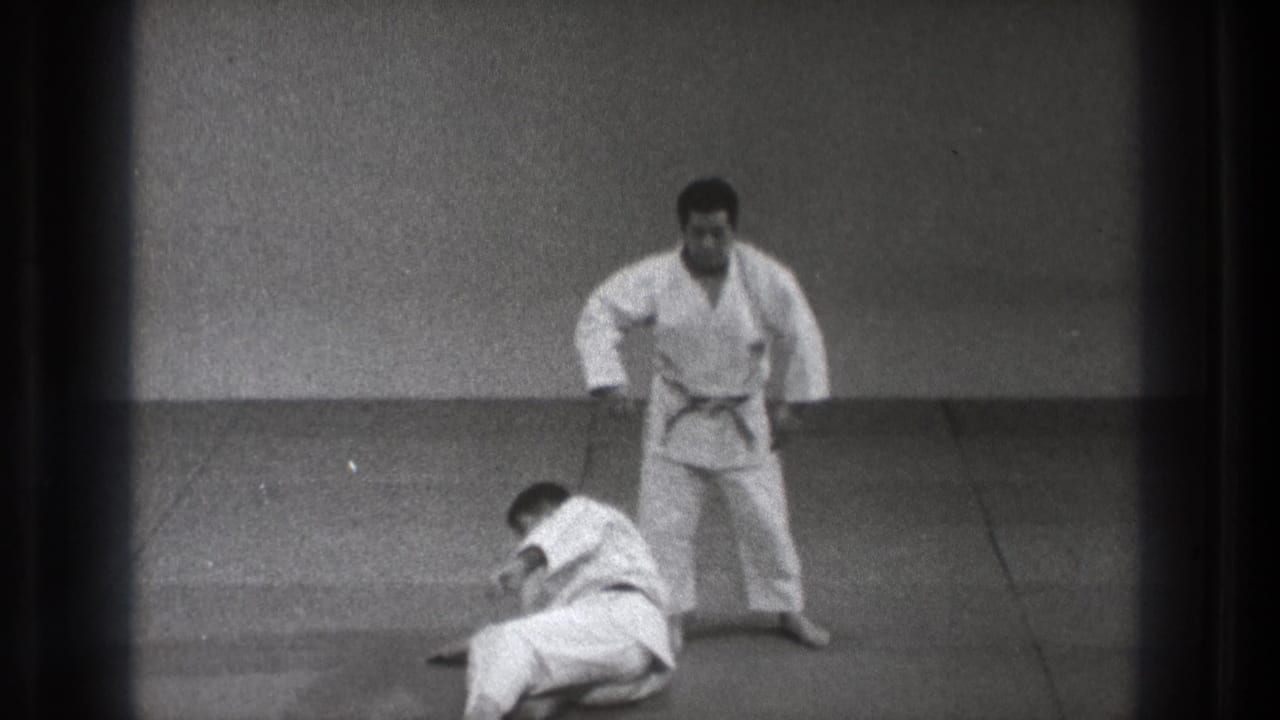 At the end of the 1971 Tokyo judo sequence, one man stands beside his partner as the thrown participant settles on the mat.