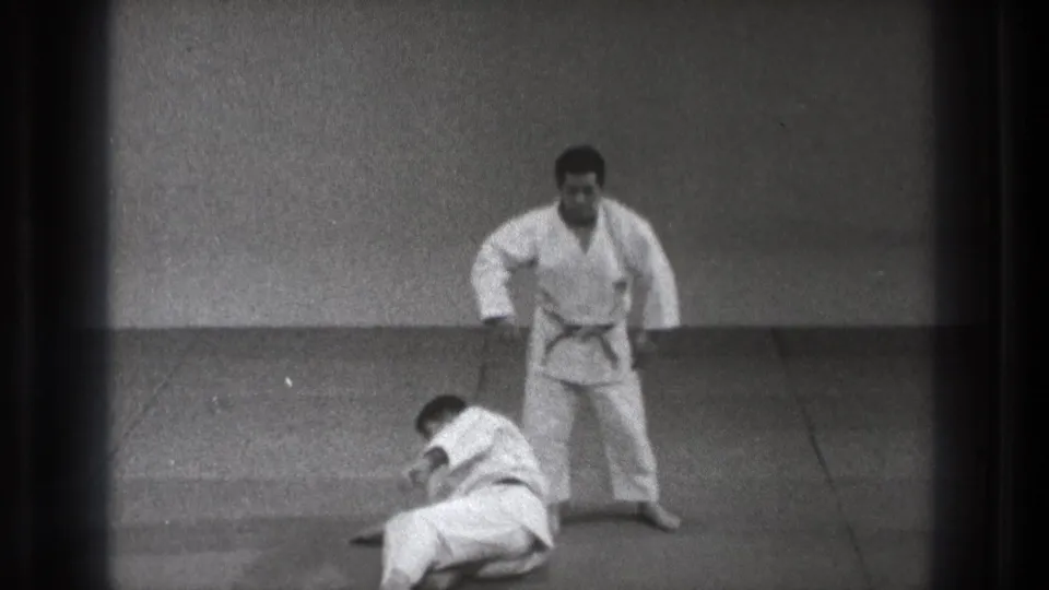 At the end of the 1971 Tokyo judo sequence, one man stands beside his partner as the thrown participant settles on the mat.