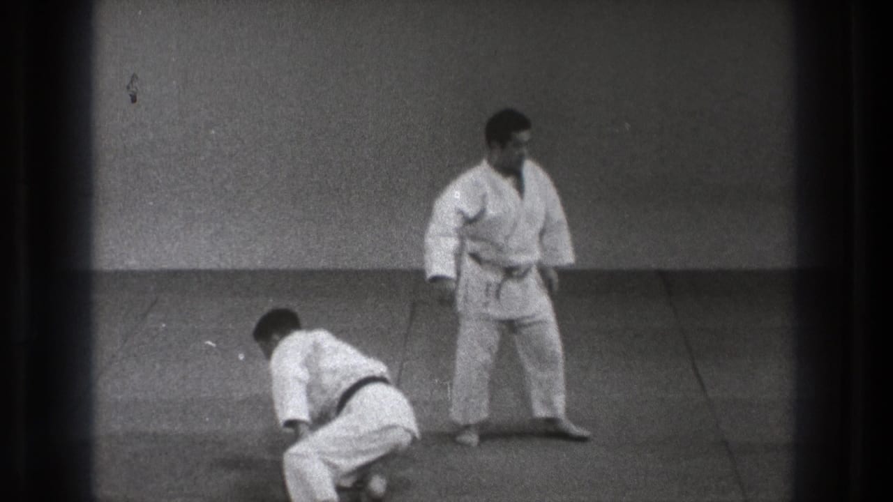 Two men in white martial arts uniforms on a mat, one kneeling and one standing.