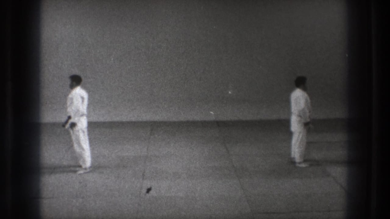 Two martial artists in white gi stand apart on a plain mat floor.