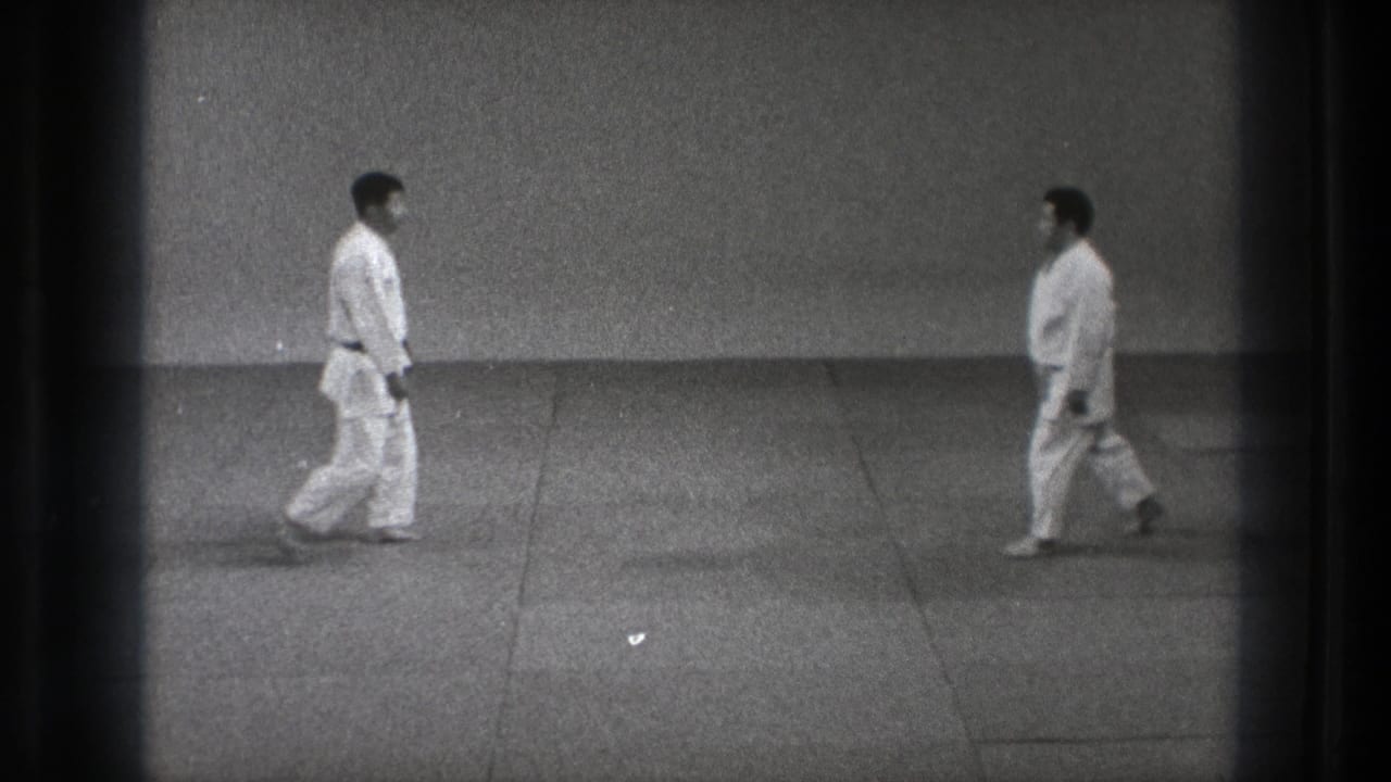 Two adult practitioners in white gi walk toward each other across a mat in black-and-white 8mm footage from the supplied Tokyo 1971 clip.