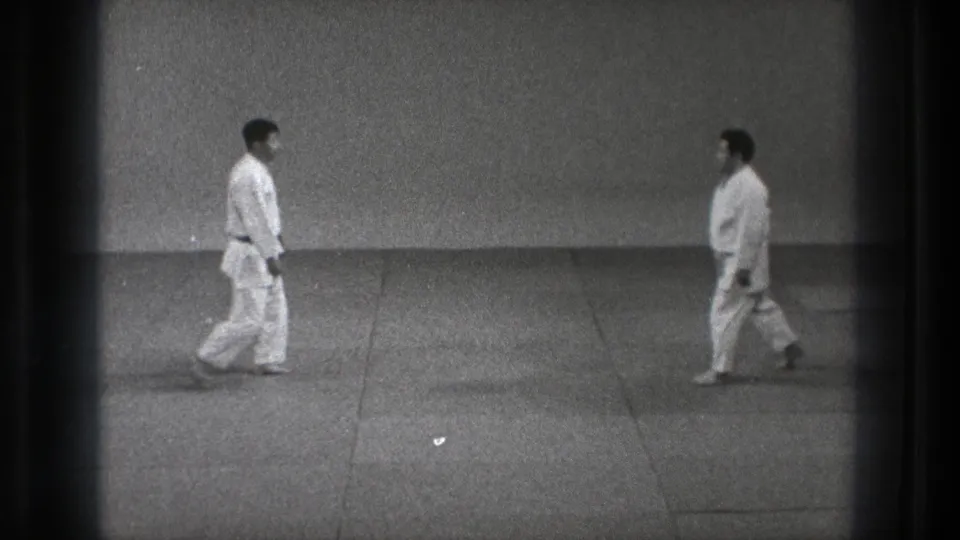 Two adult practitioners in white gi walk toward each other across a mat in black-and-white 8mm footage from the supplied Tokyo 1971 clip.