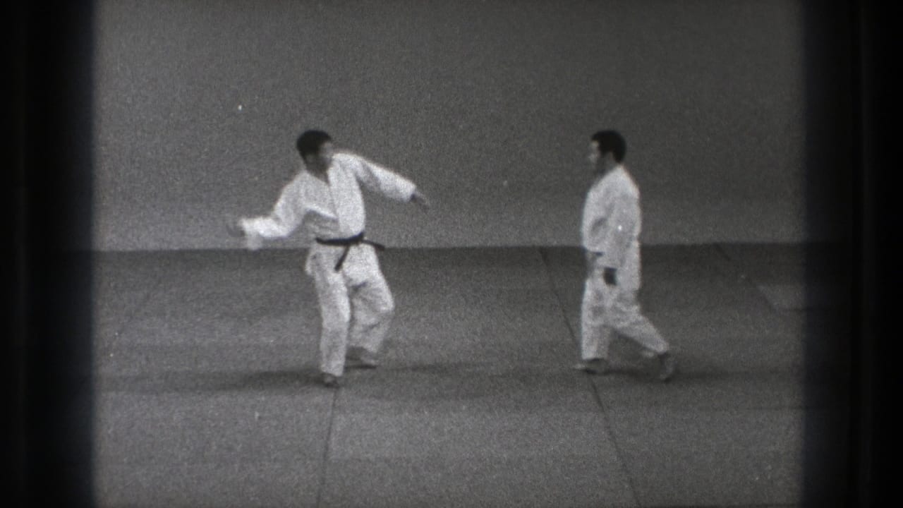 Two men in white gi face each other on floor mats in a plain indoor training room.