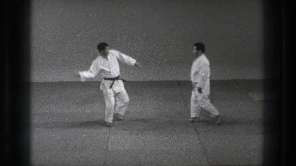In 1971 Tokyo footage, two adult karate practitioners in white gi face each other on mats, one stepping into a stance.