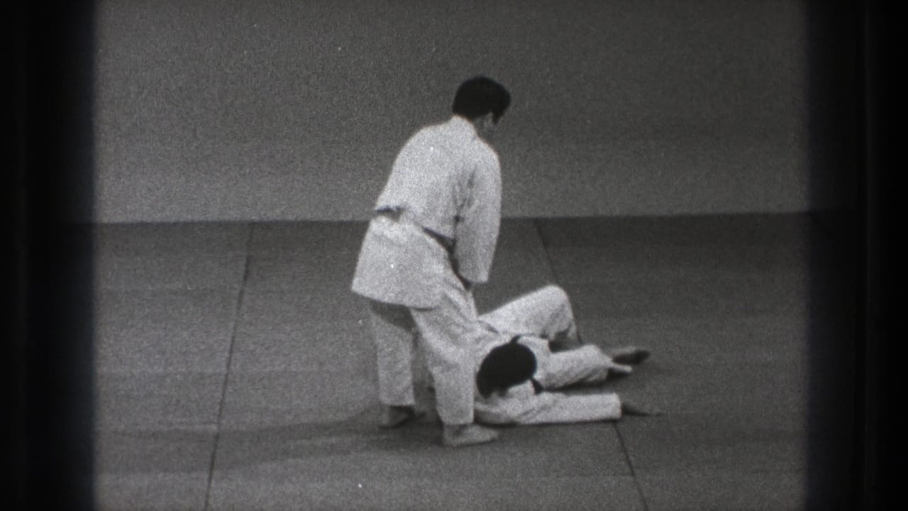 After the takedown, one karate practitioner stands near the legs of the other, who lies on the padded floor in the 1971 Tokyo footage.