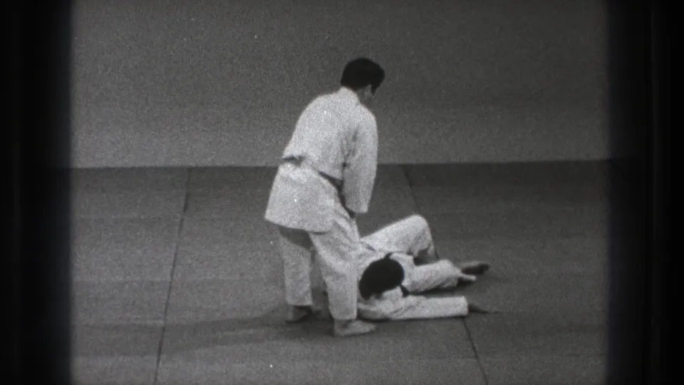 After the takedown, one karate practitioner stands near the legs of the other, who lies on the padded floor in the 1971 Tokyo footage.