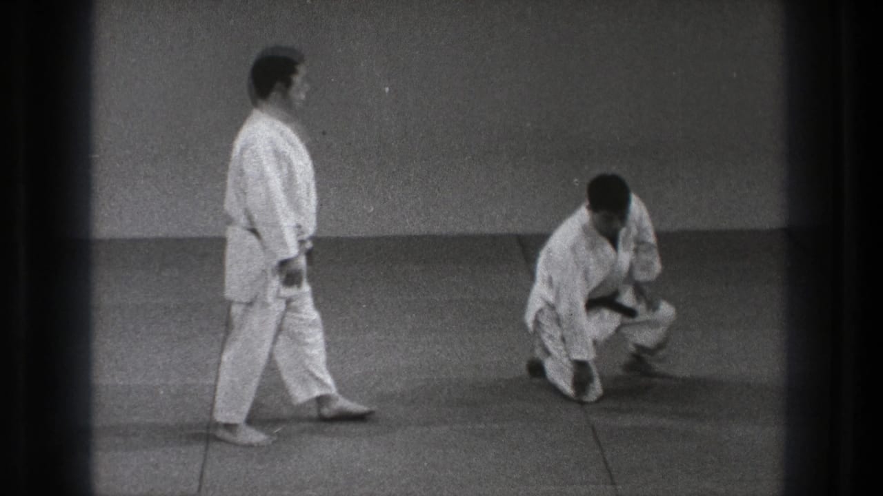 Two men in white martial arts uniforms on mats, one standing and one crouched low.