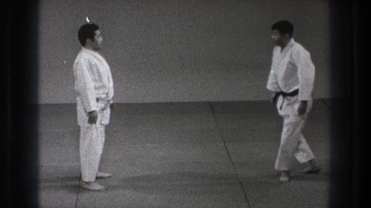 The two adult martial artists face each other in formal readiness on a mat, with one wearing a black belt in the Tokyo home-movie footage.
