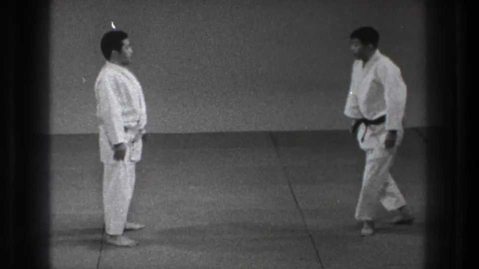 The two adult martial artists face each other in formal readiness on a mat, with one wearing a black belt in the Tokyo home-movie footage.