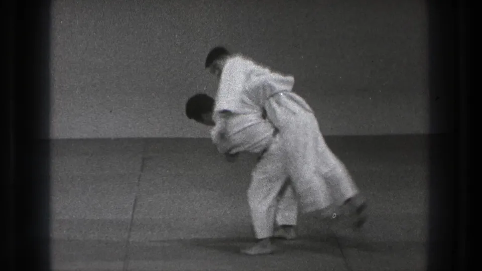 The martial artists are locked in a close throw or takedown movement on the dojo mat in this black-and-white 8mm frame.