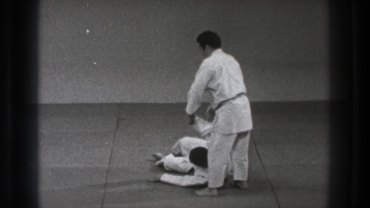 A judo practitioner in a white gi stands over a partner on the mat in the supplied 1971 Tokyo home-movie footage.