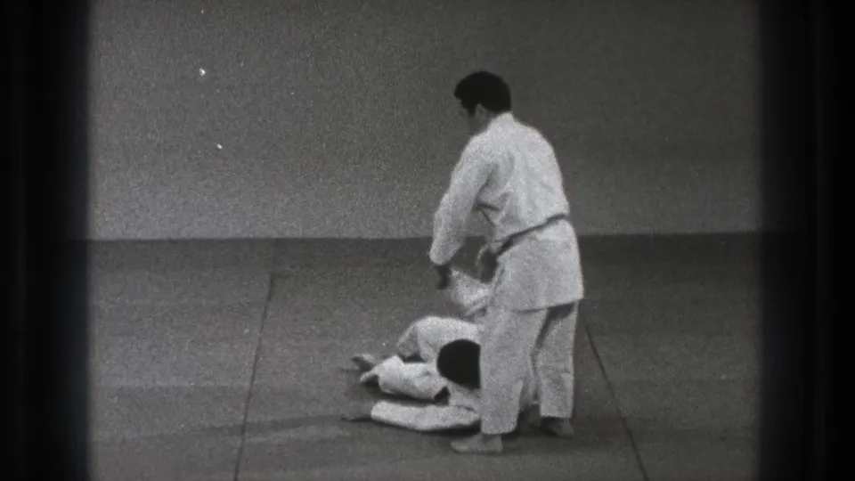 A judo practitioner in a white gi stands over a partner on the mat in the supplied 1971 Tokyo home-movie footage.