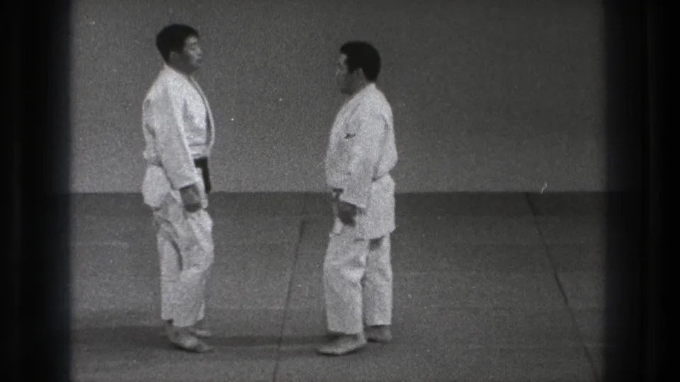 Two adult judoka in white gi and dark belts face each other on a dojo mat in Tokyo, 1971 footage.