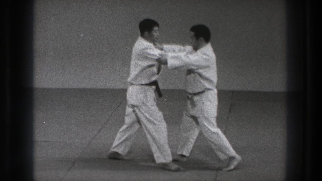 Two judoka in white gi grip each other on a dojo mat during a controlled judo exchange in 1971 Tokyo footage.