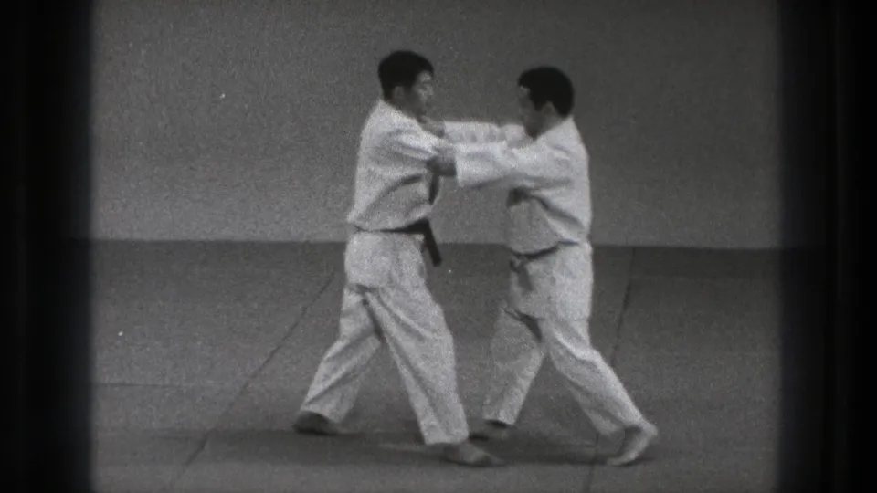 Two judoka in white gi grip each other on a dojo mat during a controlled judo exchange in 1971 Tokyo footage.