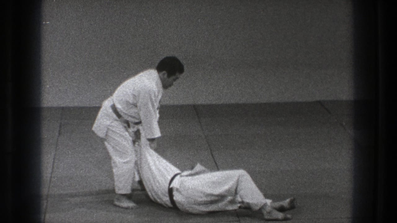 After the throw, the black-belt practitioner stands over his partner, who has landed on the mat in the Tokyo practice sequence.