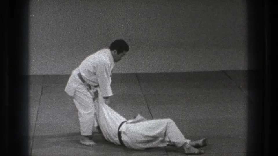 After the throw, the black-belt practitioner stands over his partner, who has landed on the mat in the Tokyo practice sequence.