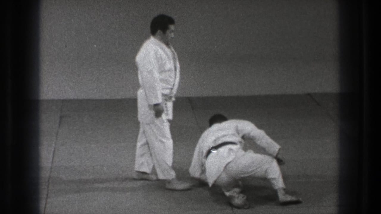A martial arts practitioner in a white gi stands nearby as his partner rises from the mat in 1971 Tokyo home-movie footage.