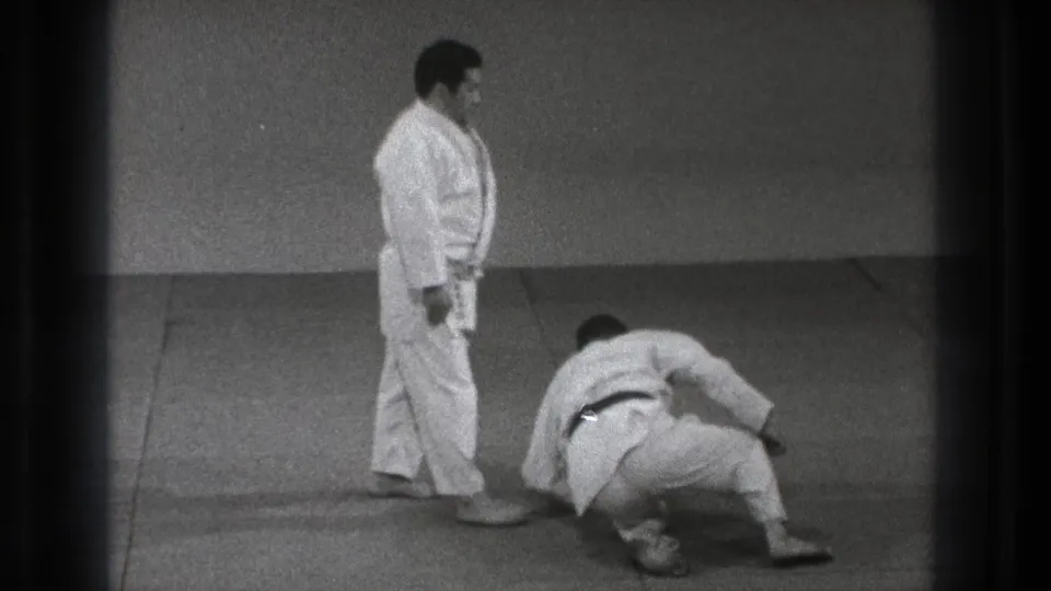 A martial arts practitioner in a white gi stands nearby as his partner rises from the mat in 1971 Tokyo home-movie footage.