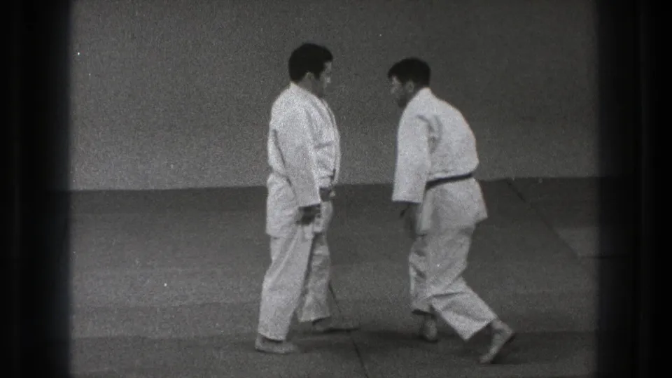 Two adult men in white gi and dark belts face each other on mats during a brief karate or martial arts training reset in Tokyo.