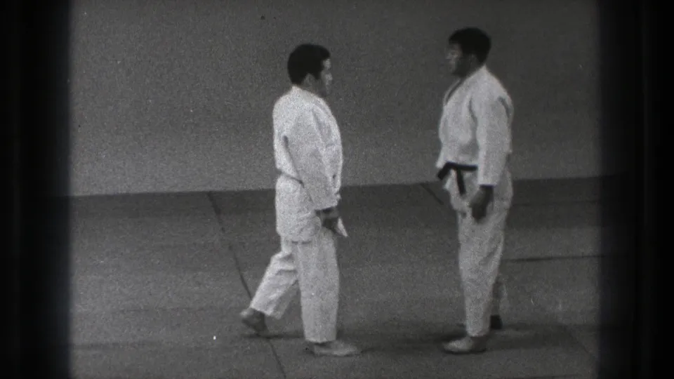 Two martial arts practitioners stand face to face on mats in a quiet training room, shown in 1971 Tokyo 8mm footage.