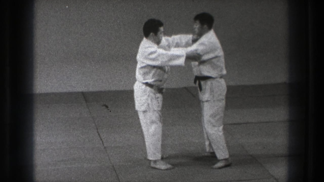 The two adult practitioners in white gi begin a defensive grab drill, with hands raised and contact at the upper body.