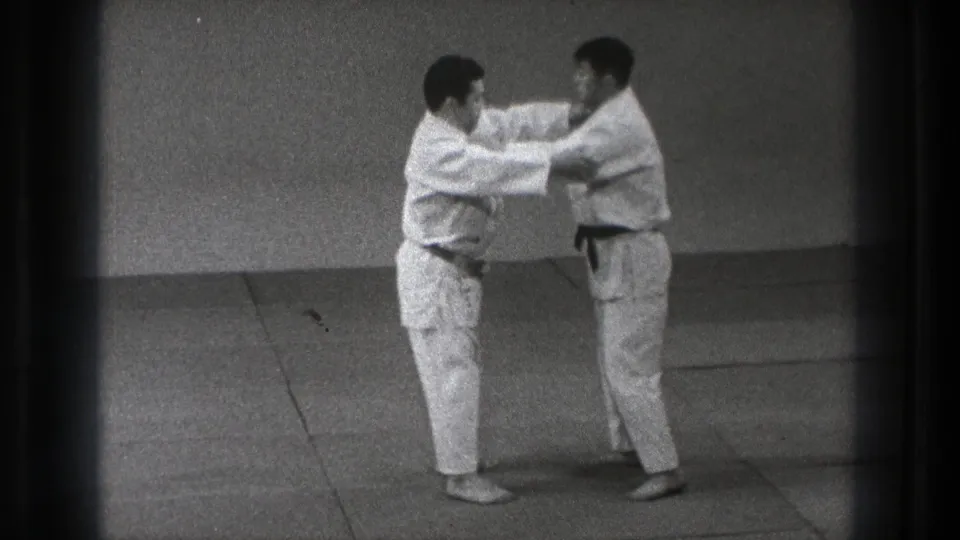The two adult practitioners in white gi begin a defensive grab drill, with hands raised and contact at the upper body.