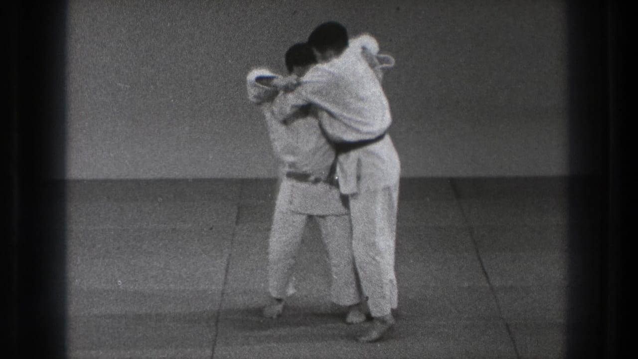 Two martial artists in white gis grip each other on an indoor mat.