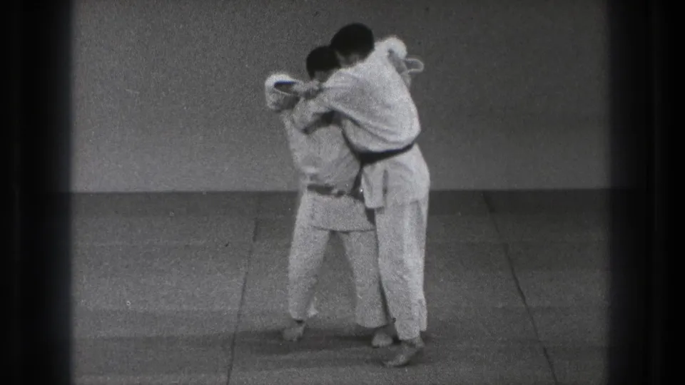 In 1971 Tokyo footage, two adult judo practitioners in white uniforms begin a throw setup on a plain indoor mat.