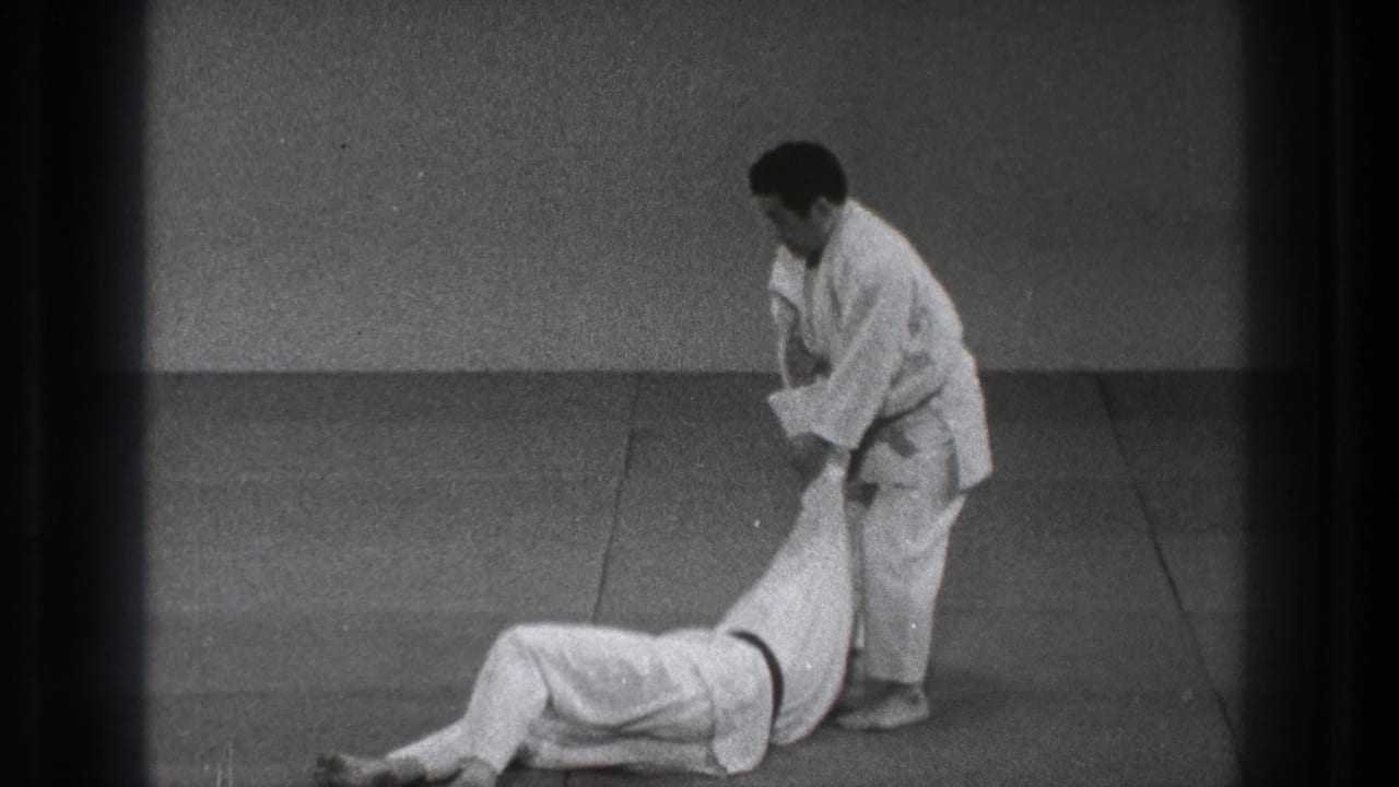 After the throw, one adult practitioner stands while holding the arm of the other, who is lying back on the mat.