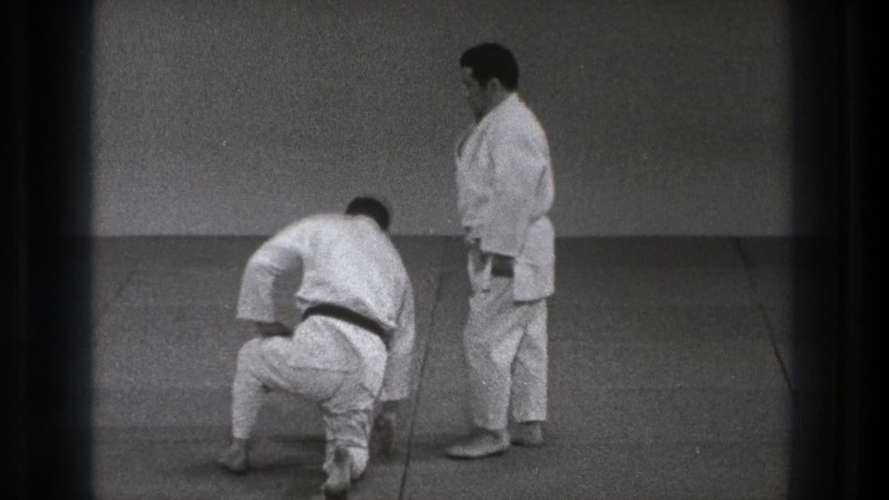 Two men in white gi on a mat, one kneeling while the other stands nearby.