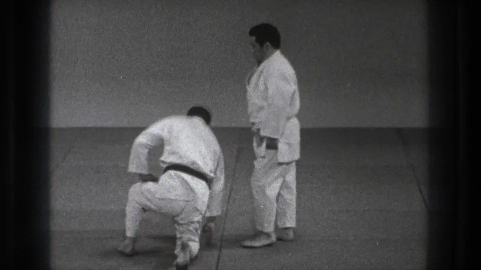 In supported Tokyo 1971 8mm footage, two adult martial artists in white gi are on a mat, with one rising from a kneeling position.