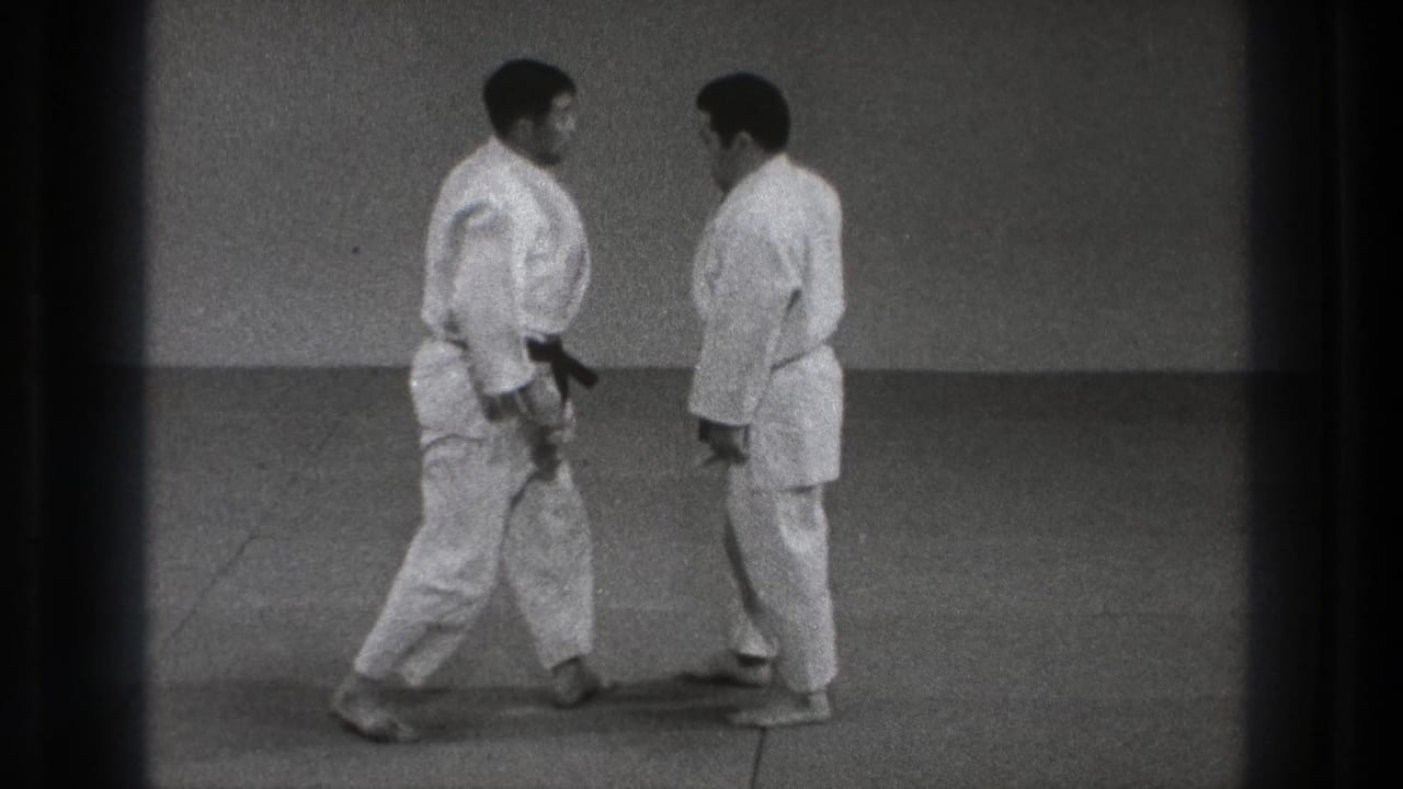 Two adult martial artists in white gi face each other on a mat in the supplied Tokyo 1971 judo practice footage.