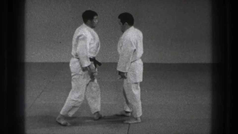Two adult martial artists in white gi face each other on a mat in the supplied Tokyo 1971 judo practice footage.