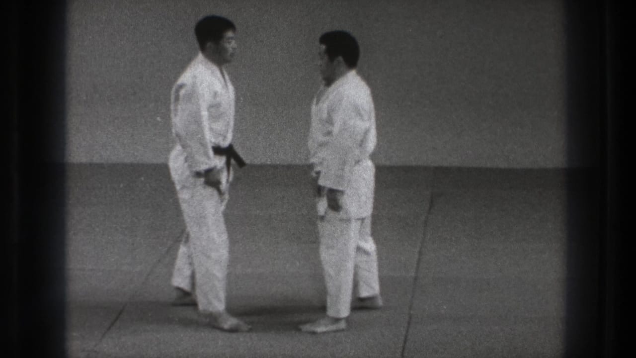 In this 1971 Tokyo martial arts frame, two adult men in white gi stand close together on a plain indoor mat area.