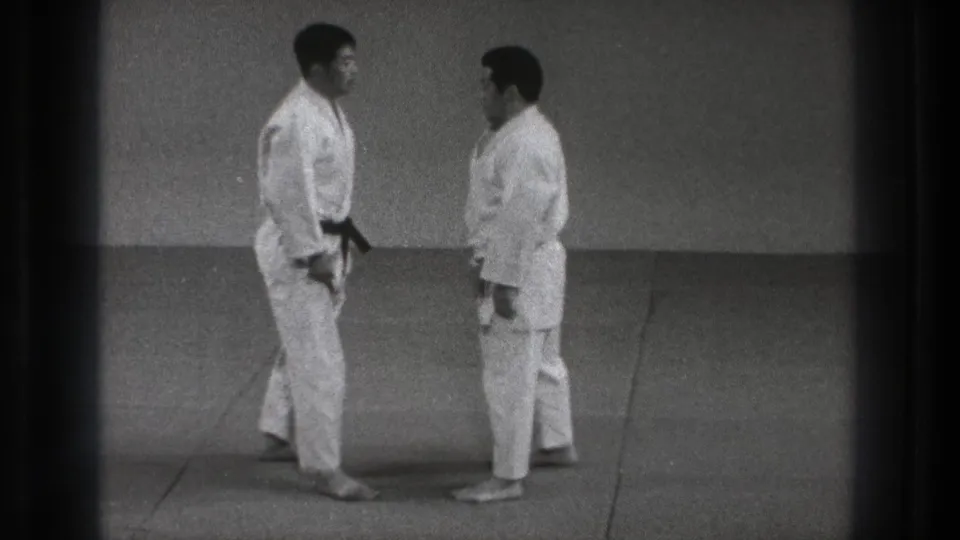 In this 1971 Tokyo martial arts frame, two adult men in white gi stand close together on a plain indoor mat area.