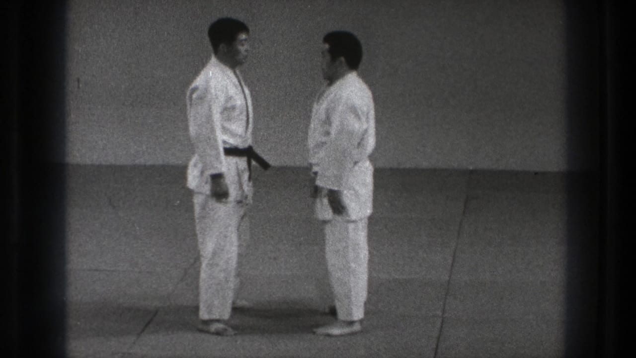 Two adult judo practitioners in white gi stand face to face on a mat in supported Tokyo 1971 home-movie footage.