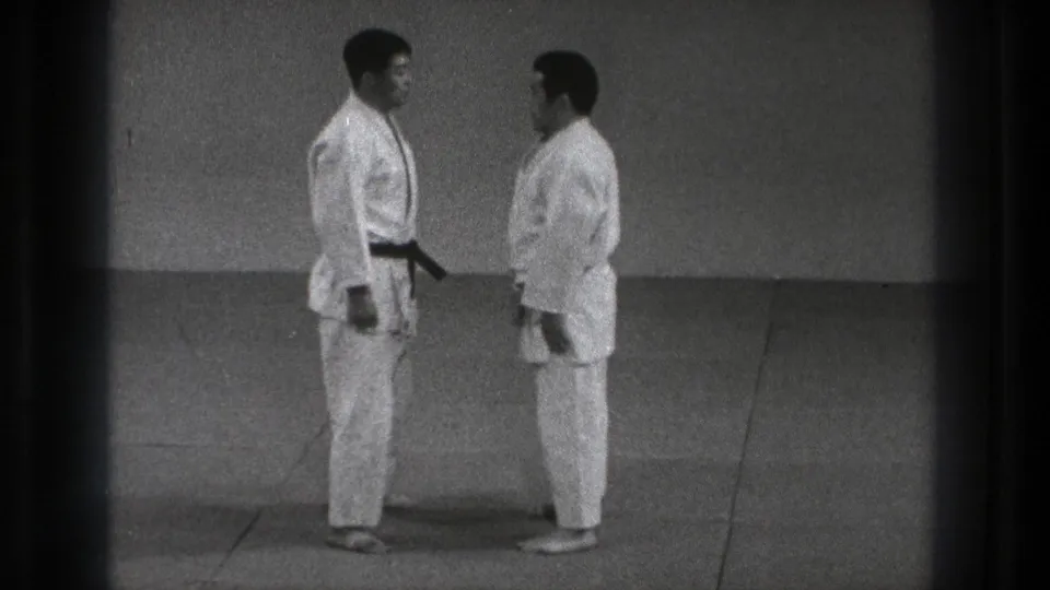 Two adult judo practitioners in white gi stand face to face on a mat in supported Tokyo 1971 home-movie footage.