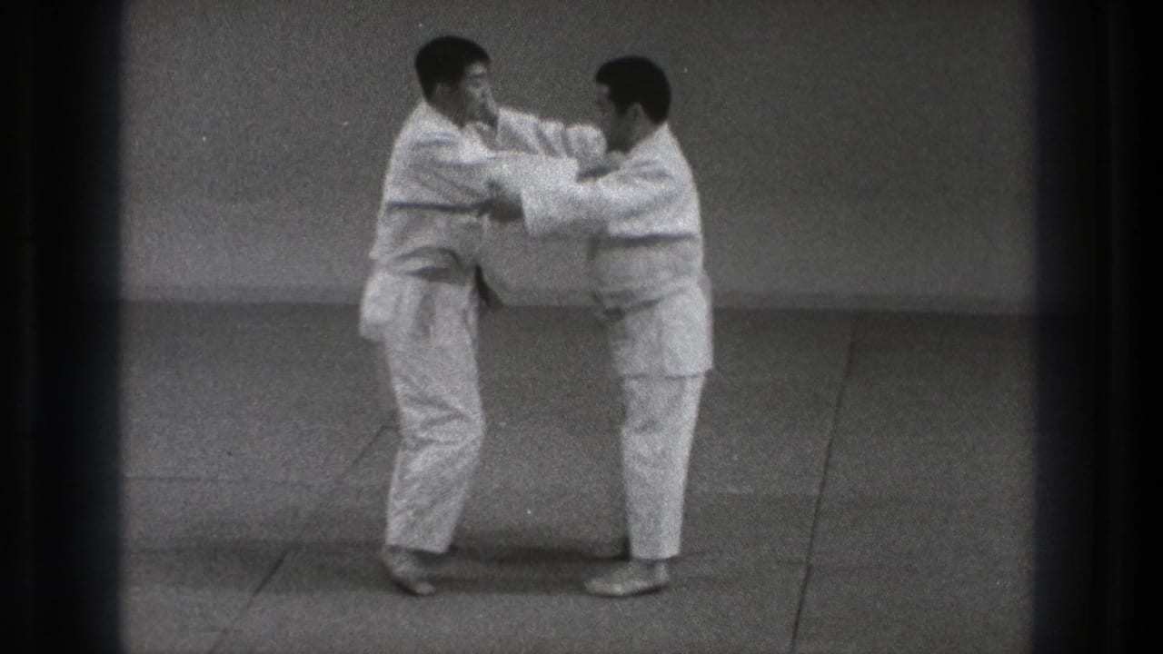 Two men in karate gi face each other on an indoor mat while gripping arms.
