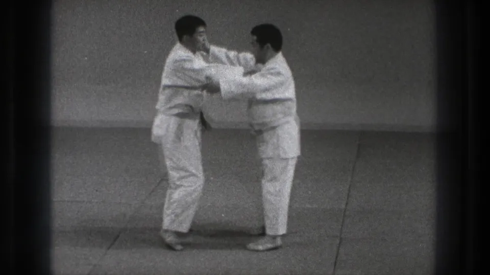 In 1971 Tokyo footage, two adult men in white karate gi stand on a plain indoor mat during a paired technique drill.