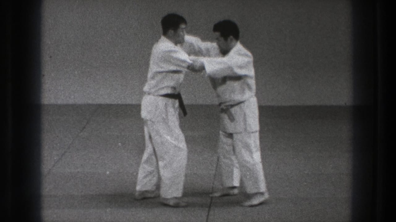 Two adult practitioners in white gi continue a controlled karate defense movement on an indoor mat in the Tokyo home-movie clip.