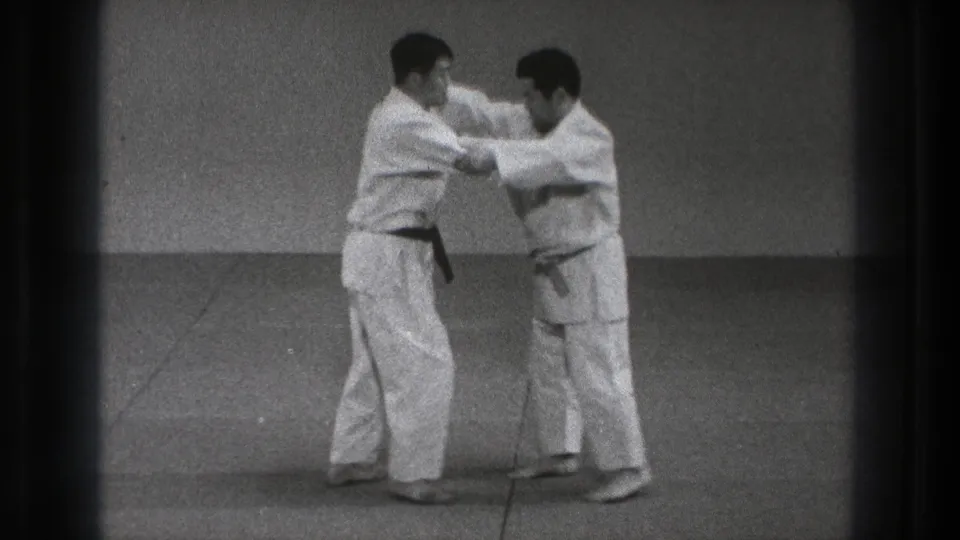 Two adult practitioners in white gi continue a controlled karate defense movement on an indoor mat in the Tokyo home-movie clip.